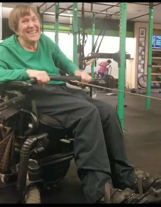 A laughing athlete in a power wheelchair pulls a rowing handle during an adaptive cable workout