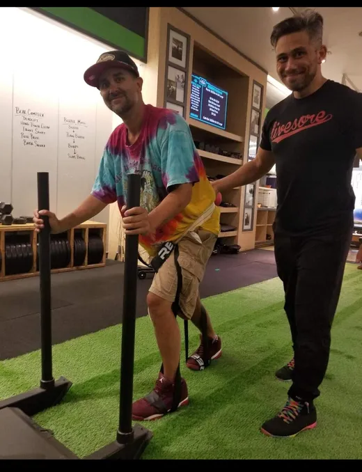 An athlete pushes a weighted sled across the turf while trainer Sean walks alongside him