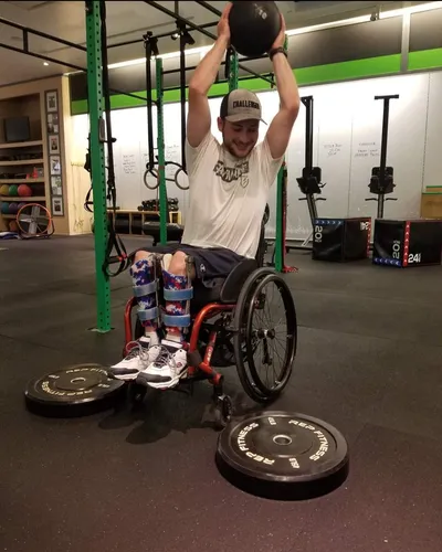 A smiling adaptive athlete wearing leg braces lifts a medicine ball overhead from his wheelchair, with bumper plates on the floor beside him