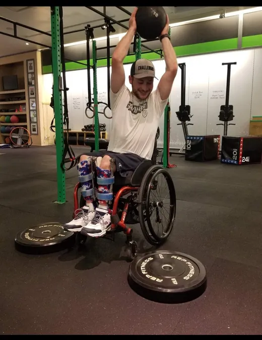 A smiling adaptive athlete wearing leg braces lifts a medicine ball overhead from his wheelchair, with bumper plates on the floor beside him